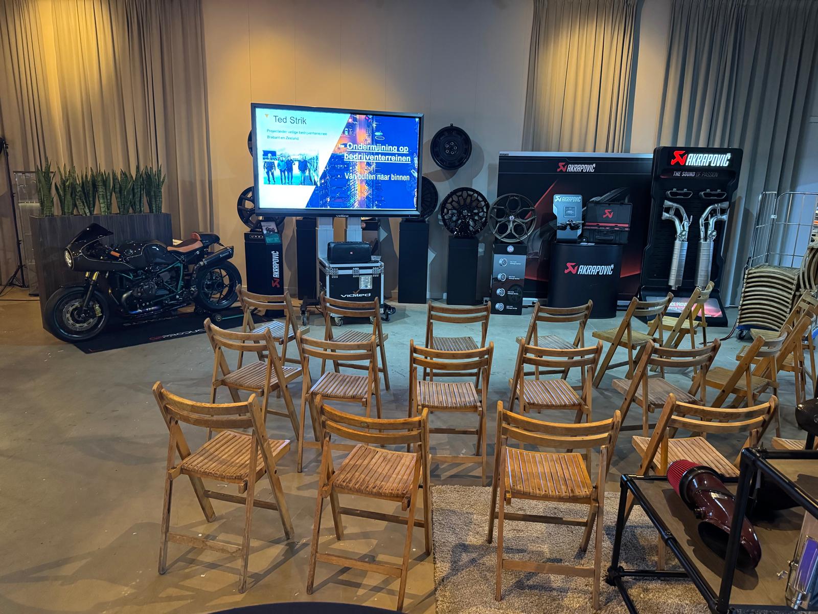 Overzicht van de presentatie-opstelling met lege houten stoelen gericht op een groot mobiel beeldscherm op een flightcase. Rondom de opstelling staan exclusieve auto- en motoronderdelen tentoongesteld.