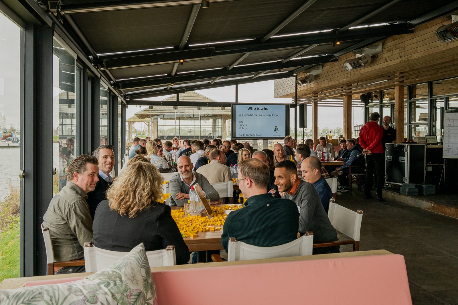 Zijaanzicht van een drukke workshop bij More-Itz, waar deelnemers aan tafels zitten te kijken naar een LCD-scherm met de presentatietitel "Wie is wie" en een spreker in een rode trui die uitleg geeft bij een tafel met gele LEGO-stenen.