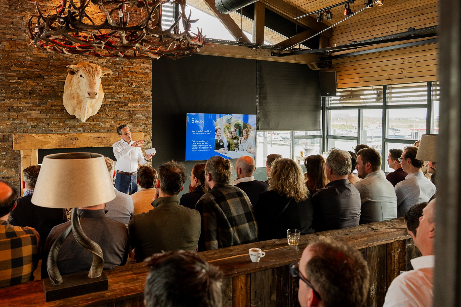 Achteraanzicht van het publiek tijdens de presentatie van Studibo bij More-Itz. De spreker staat naast een grote stenen open haard en een opgezet dier, en het scherm toont een dia over evenemententypen.
