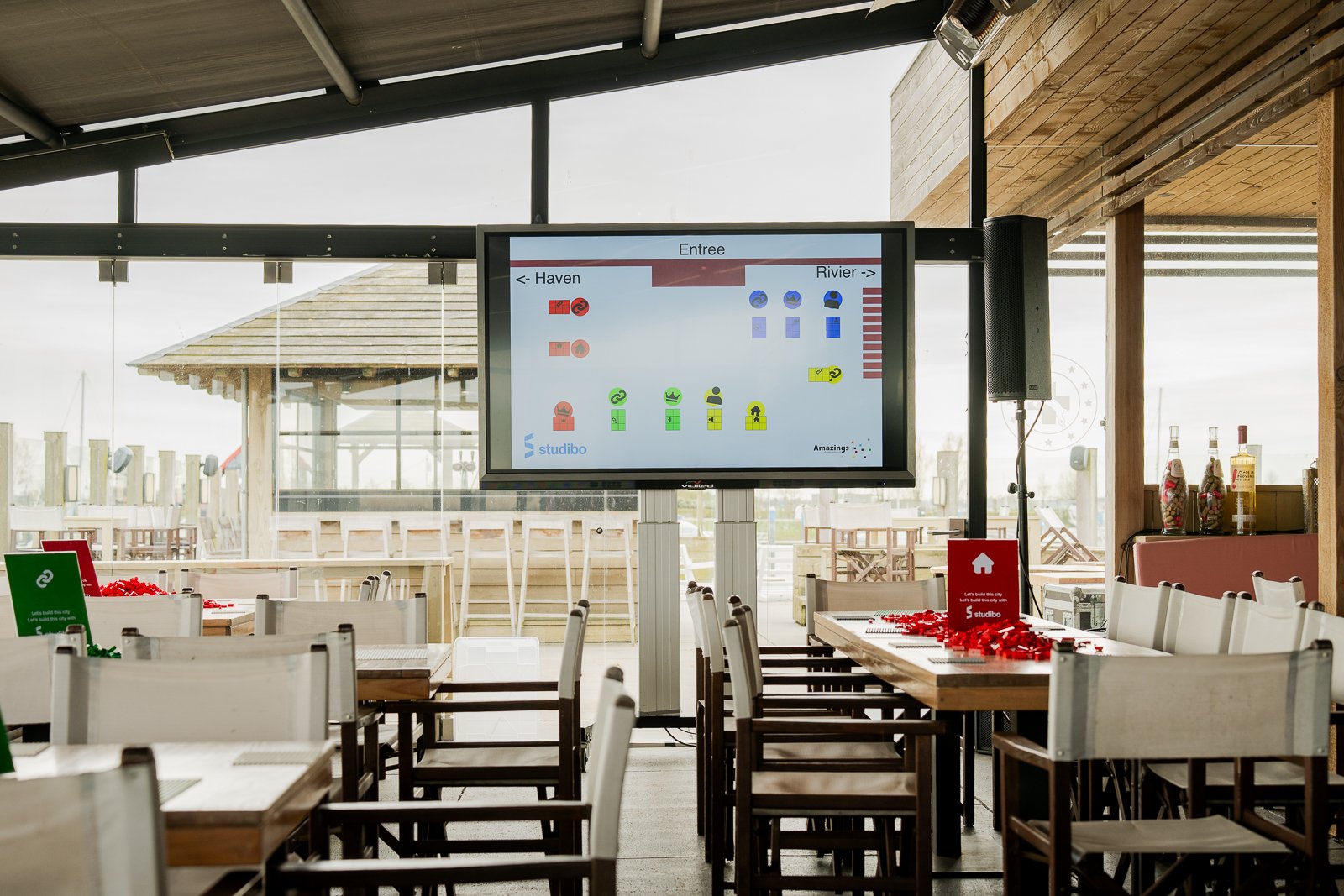Close-up van het hoofdscherm en tafels tijdens de workshop bij More-Itz. Het scherm toont een diagram van een LEGO 'Studibo' lay-out. Op de voorgrond staan tafelborden en een model gebouwd met rode LEGO-stenen.