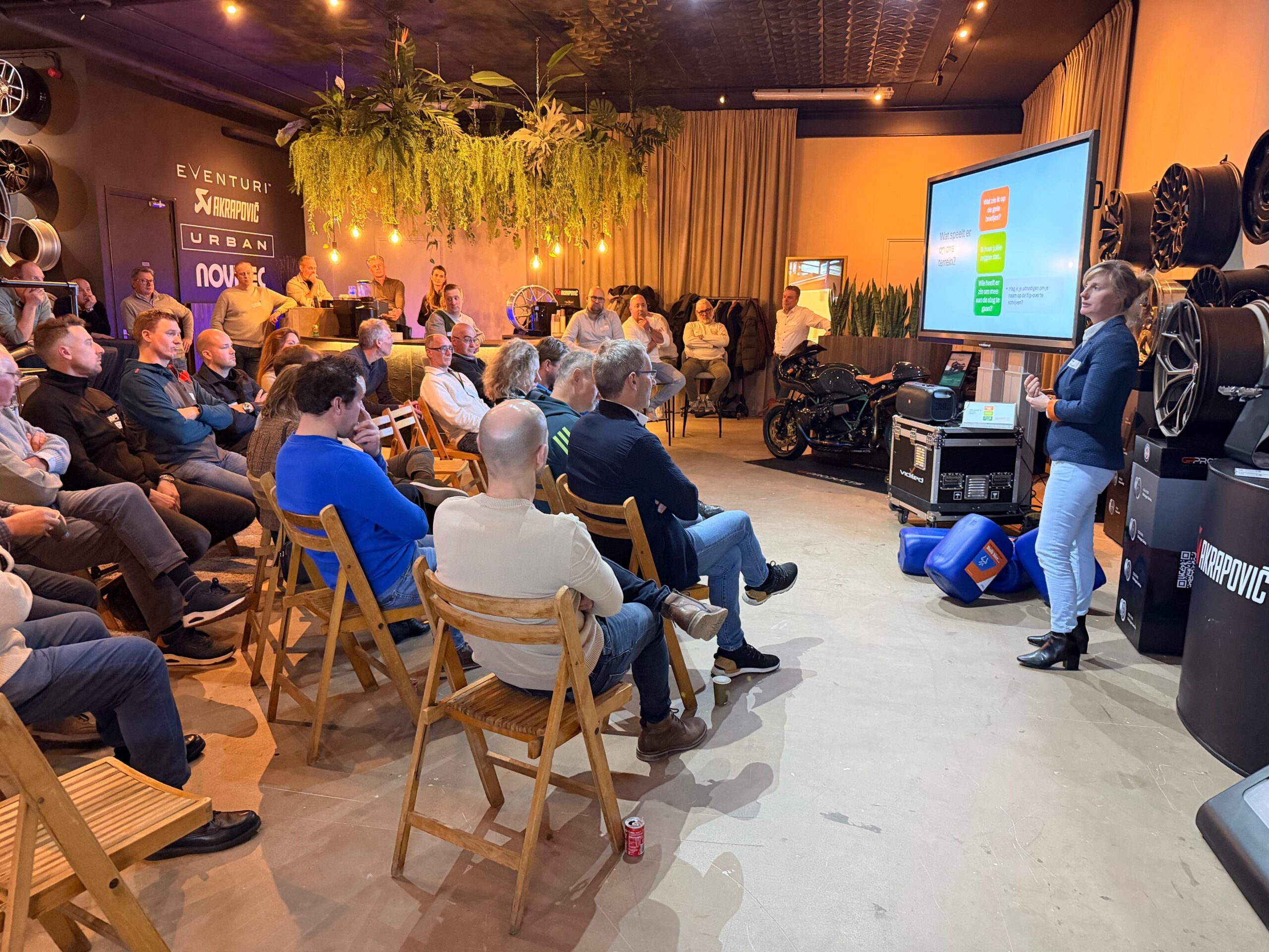 Een vrouw in een blauwe blazer geeft een presentatie voor een aandachtig zittend publiek van ondernemers in Loon op Zand, met naast haar een groot scherm en een custom motorfiets op de achtergrond.