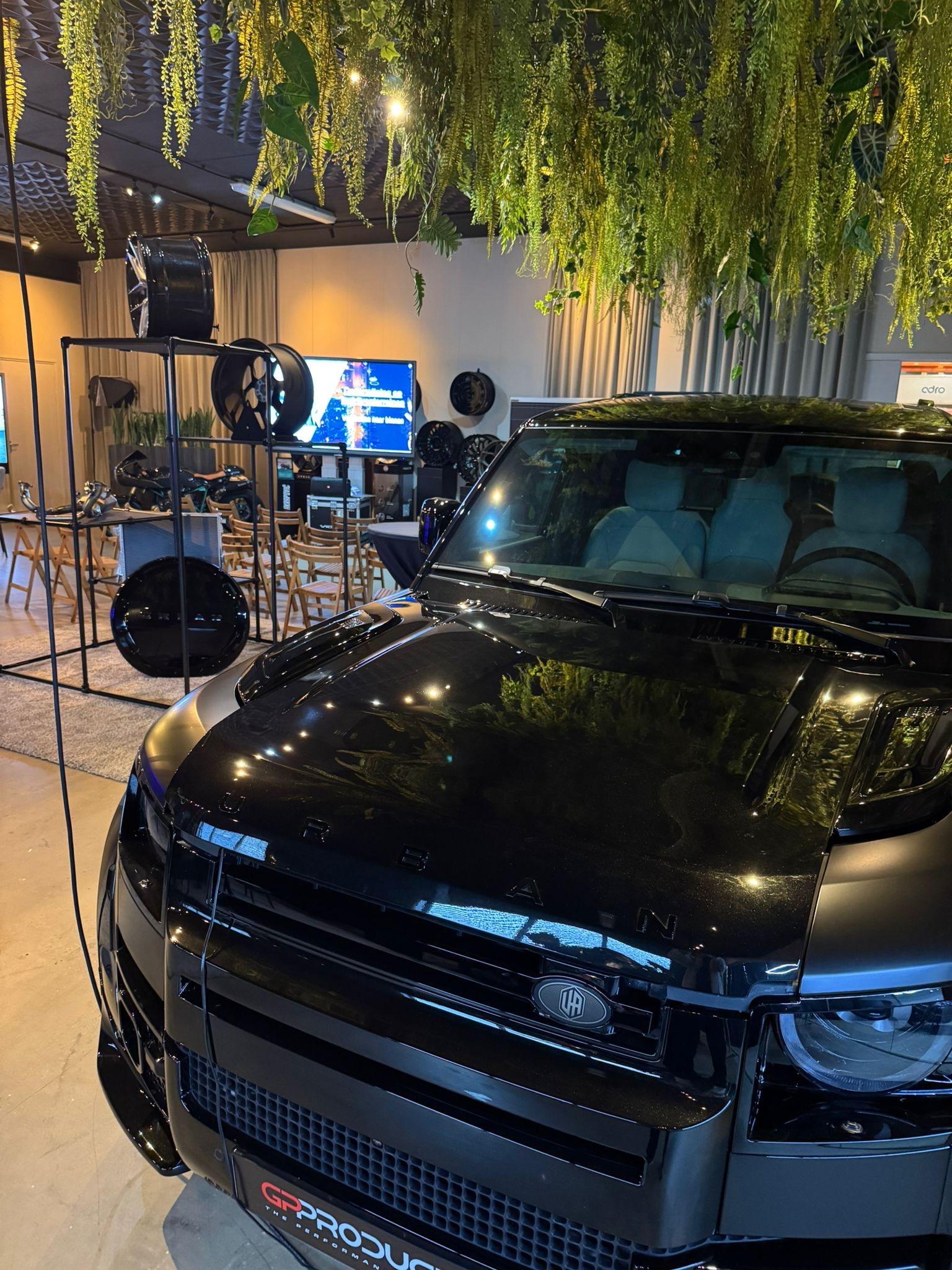 Glanzende zwarte Land Rover Defender op de voorgrond met op de achtergrond een opstelling van houten stoelen en een groot presentatiescherm voor de bijeenkomst van Stichting SOL.