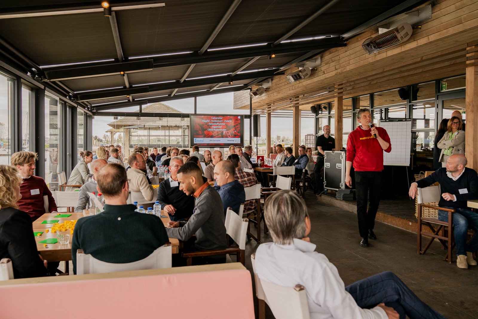 Een overzichtsfoto van de workshopruimte bij More-Itz. De spreker staat achteraan met een microfoon, en een groot publiek zit aan tafels met LEGO-materialen. Het LCD-scherm in de achtergrond toont de workshopinformatie.