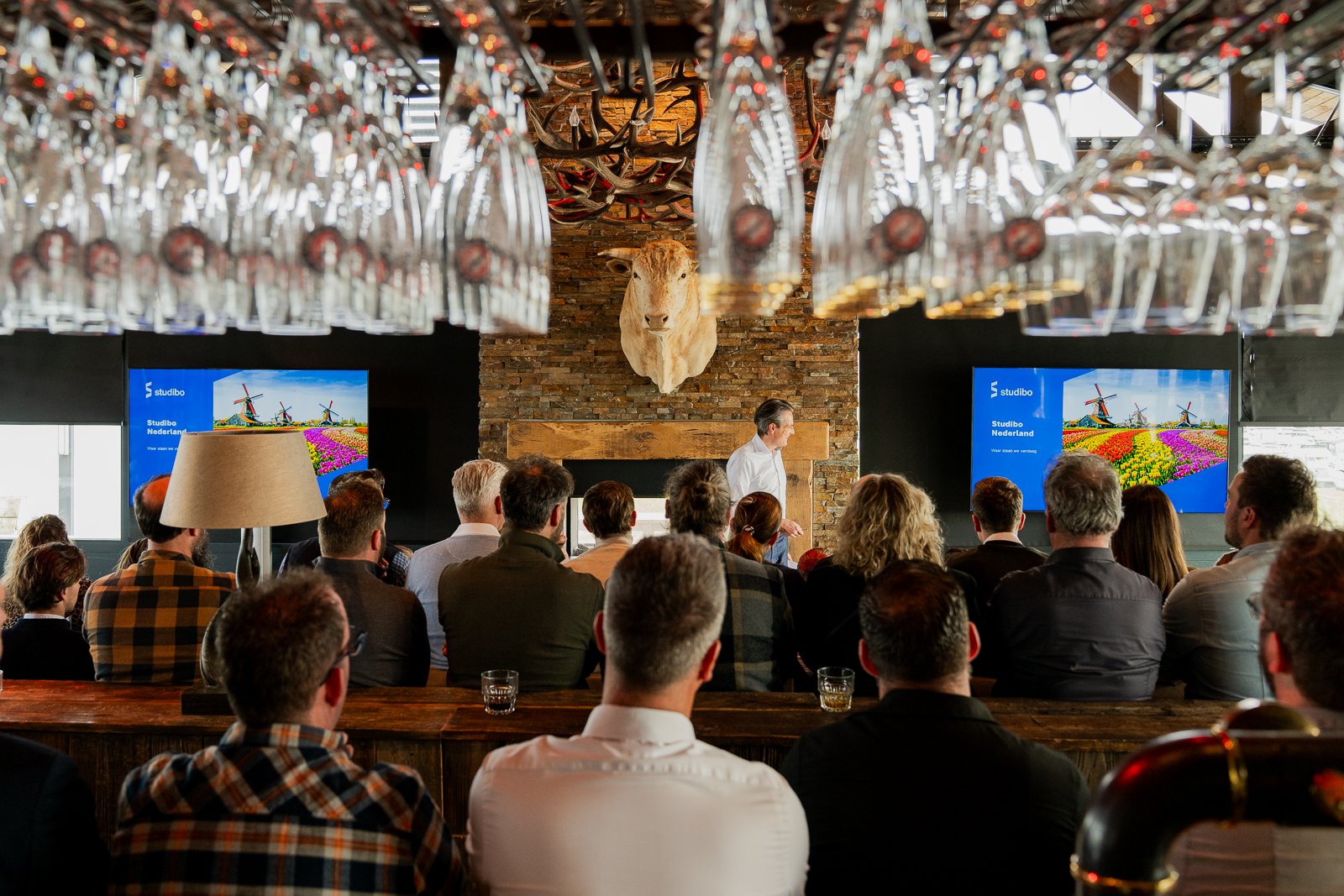 Een ingekaderd zicht op de presentatie van Studibo bij More-Itz, gezien door de hangende wijnglazen boven de bar. We kijken tegen de ruggen van het publiek aan, naar de spreker en de twee LCD-schermen met de presentatiedia bij de stenen haard.