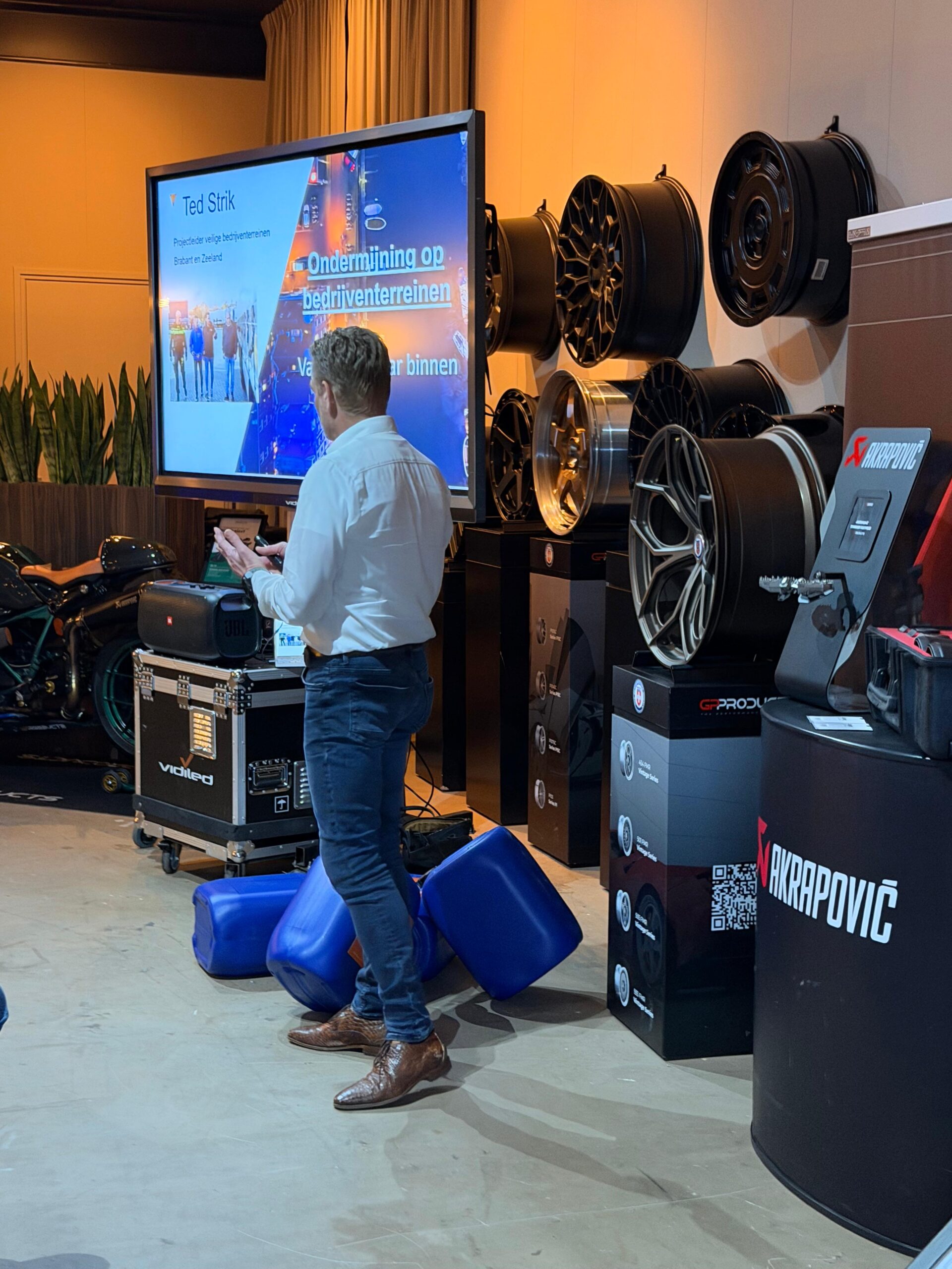 Spreker in een wit overhemd geeft een presentatie over 'Ondermijning op bedrijventerreinen' via een groot verrijdbaar beeldscherm, omringd door exclusieve autovelgen en uitlaatsystemen.