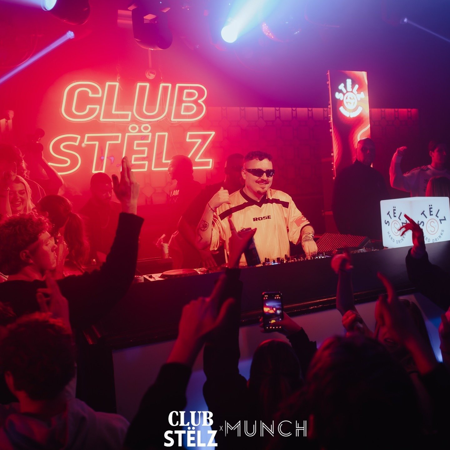 Close-up van een lachende DJ met zonnebril achter de draaitafels. Hij staat voor een opvallende rode 'CLUB STËLZ' neonwand en een verticaal LED-scherm. Op de voorgrond steken enthousiaste bezoekers hun handen in de lucht. Onderaan de foto staat het 'CLUB STËLZ x MUNCH' logo.