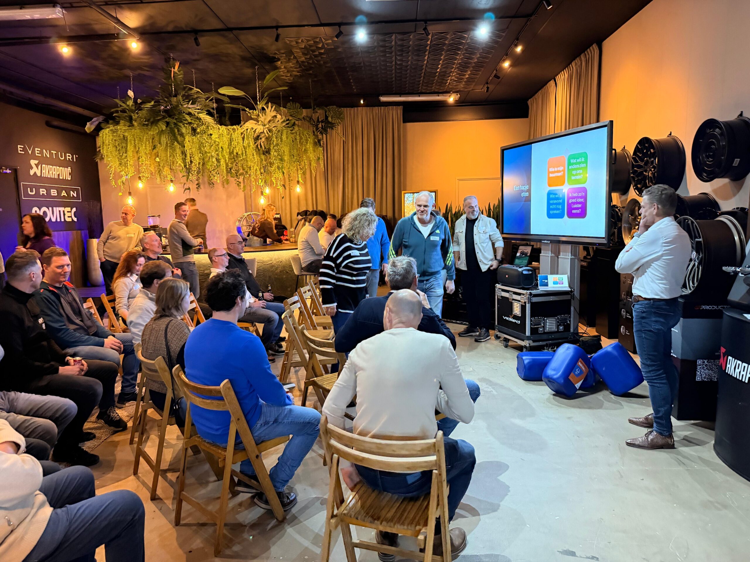 Ondernemers nemen plaats op houten klapstoeltjes tijdens een netwerkbijeenkomst en bedrijfspresentatie van Stichting Ondernemend Loon op Zand in een showroom met hangplanten en auto-onderdelen.