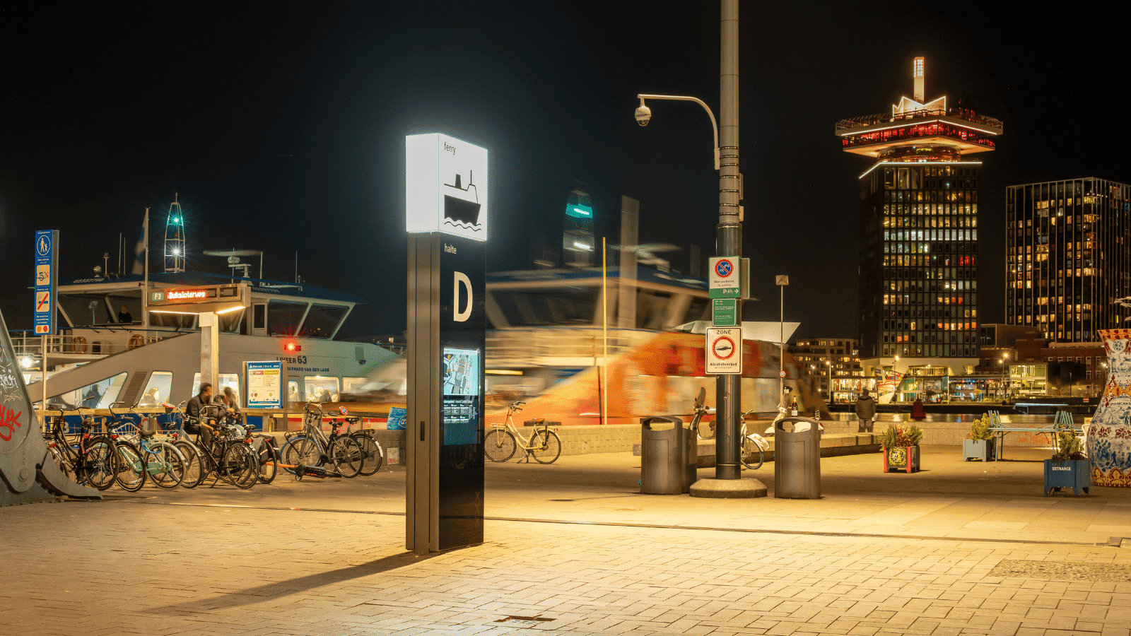 Een modern verlicht LED-wayfinding scherm bij Amsterdam Centraal Station in de avond, dat de vertrektijden van de veerponten aangeeft inclusief een kaart.